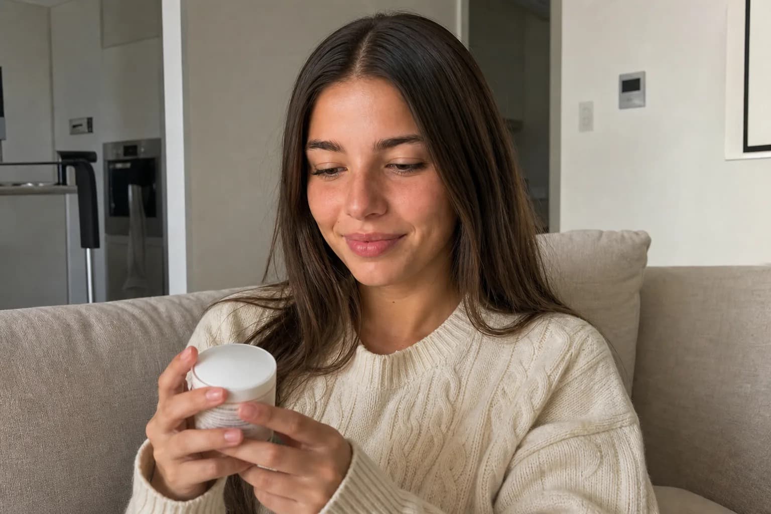 Young woman sitting on a linen sofa holding a small product jar and looking at it, afternoon light