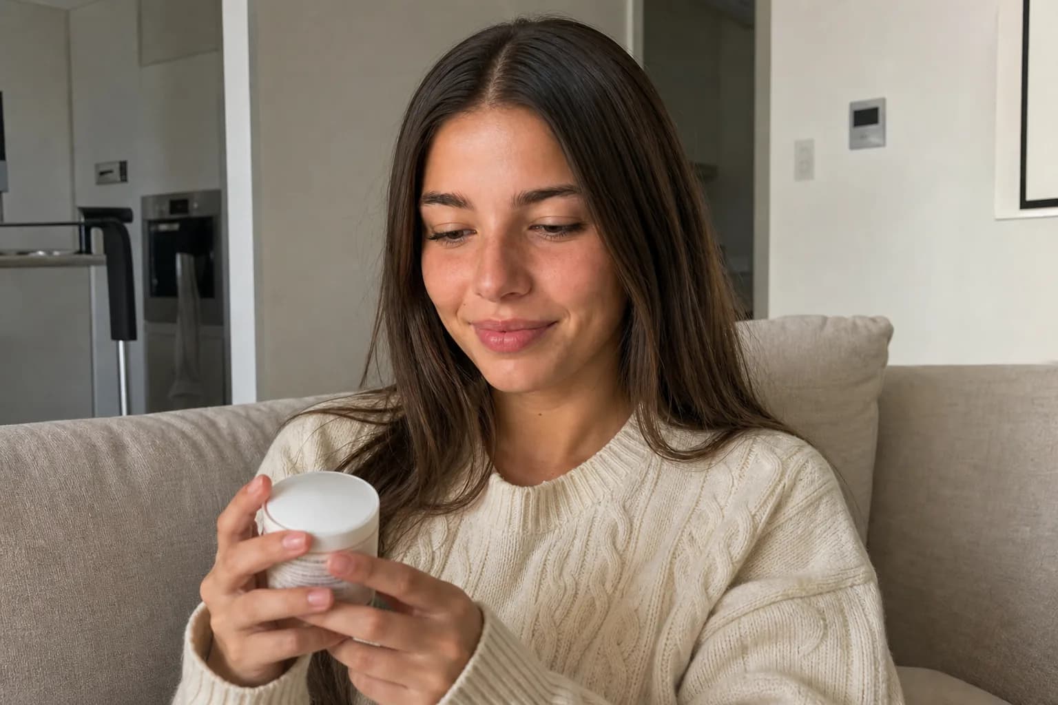Young woman sitting on a linen sofa holding a small product jar and looking at it, afternoon light