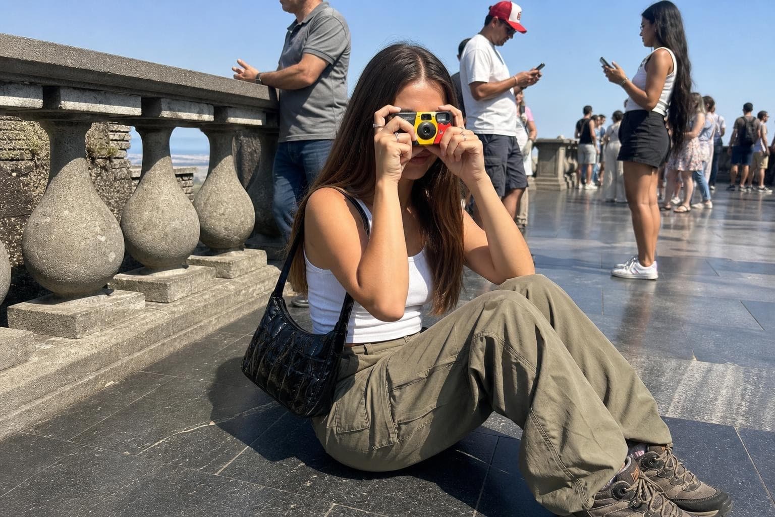 Candid AI photo of a character sitting on a crowded stone observation terrace, shooting with a compact film camera while tourists around her look at their phones, flat midday daylight and an off-center frame, the look of a real smartphone moment rather than a cinematic render