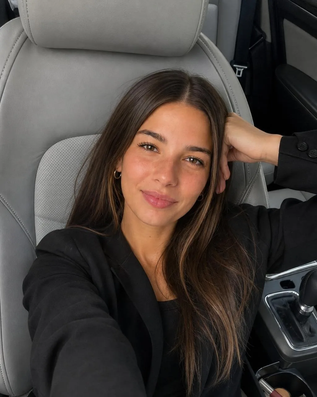 Cladegrove generated photo of the saved character taking a smiling selfie in a car seat, black blazer over a black top, soft daylight, produced from one short prompt and one credit
