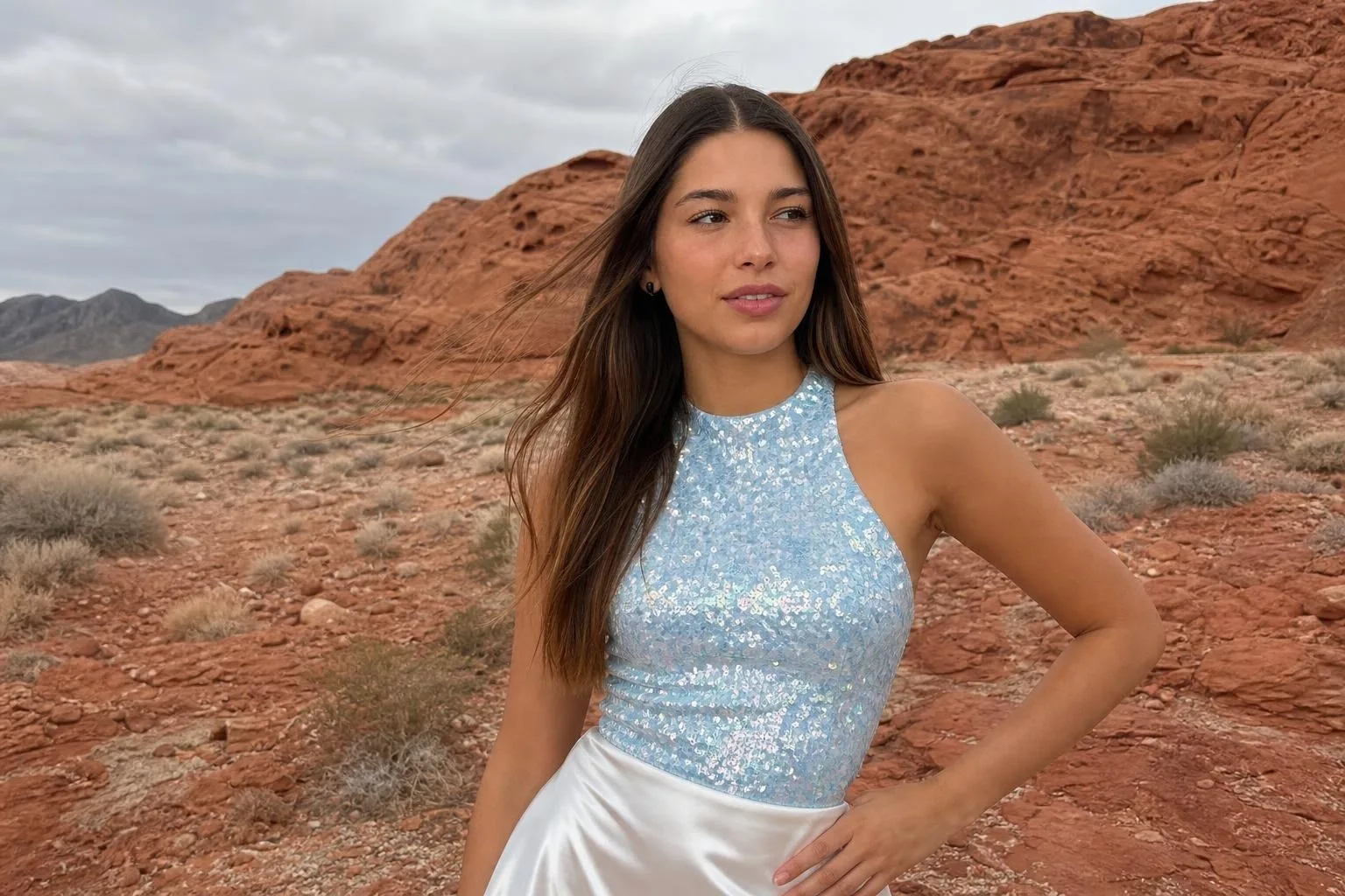 Same AI influencer in a desert of dramatic red rock formations, light-blue sequin halter top and white satin skirt, overcast sky and weathered sandstone behind her