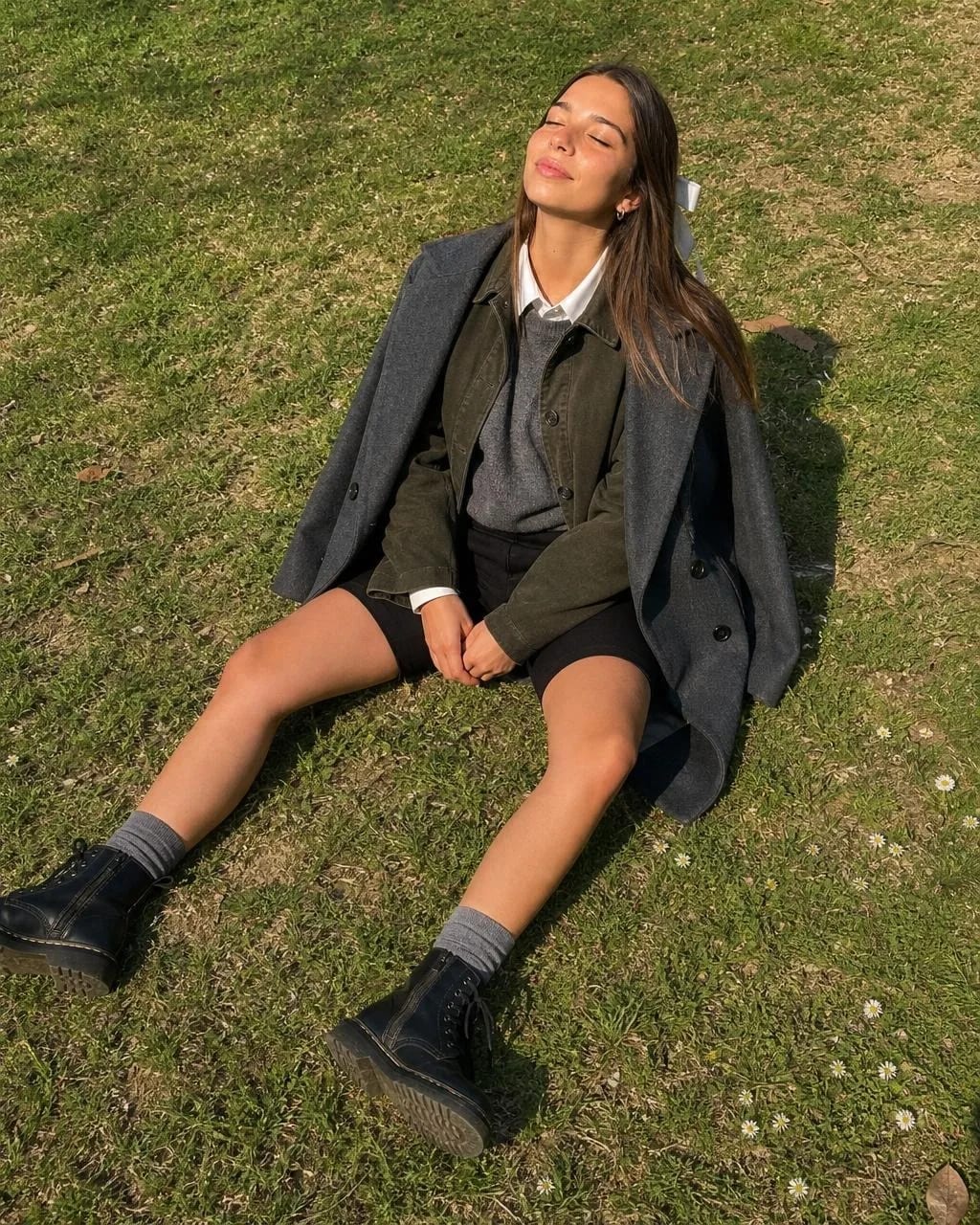 Same Cladegrove creator face sitting on sunlit grass in a public park, eyes closed toward the sun, grey wool coat over an olive jacket with black cycling shorts and chunky lace-up boots, daisies in the lawn