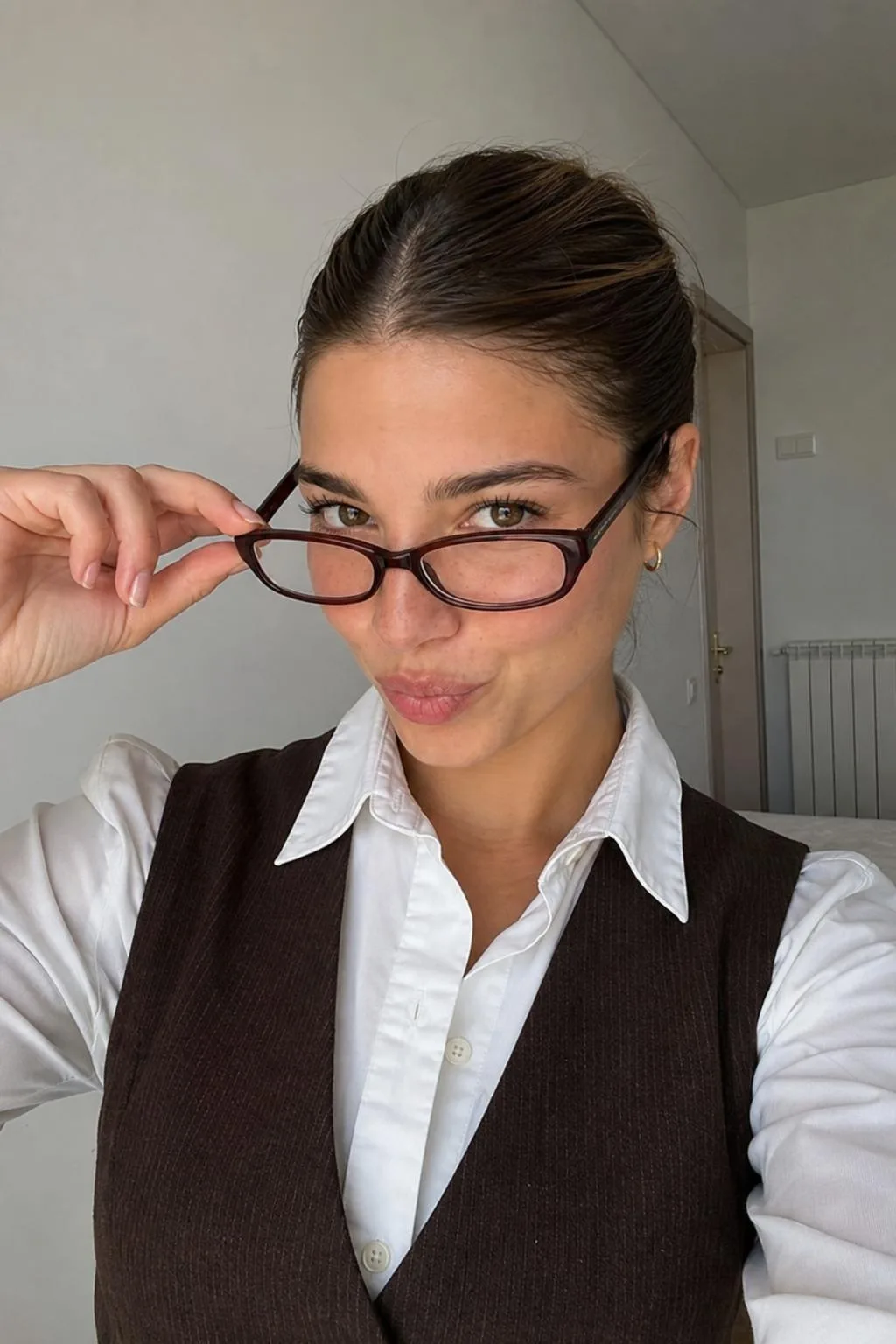 Same Cladegrove creator face in a bright minimalist room, close-up selfie lowering rectangular glasses, white shirt under a brown pinstripe vest, soft natural daylight