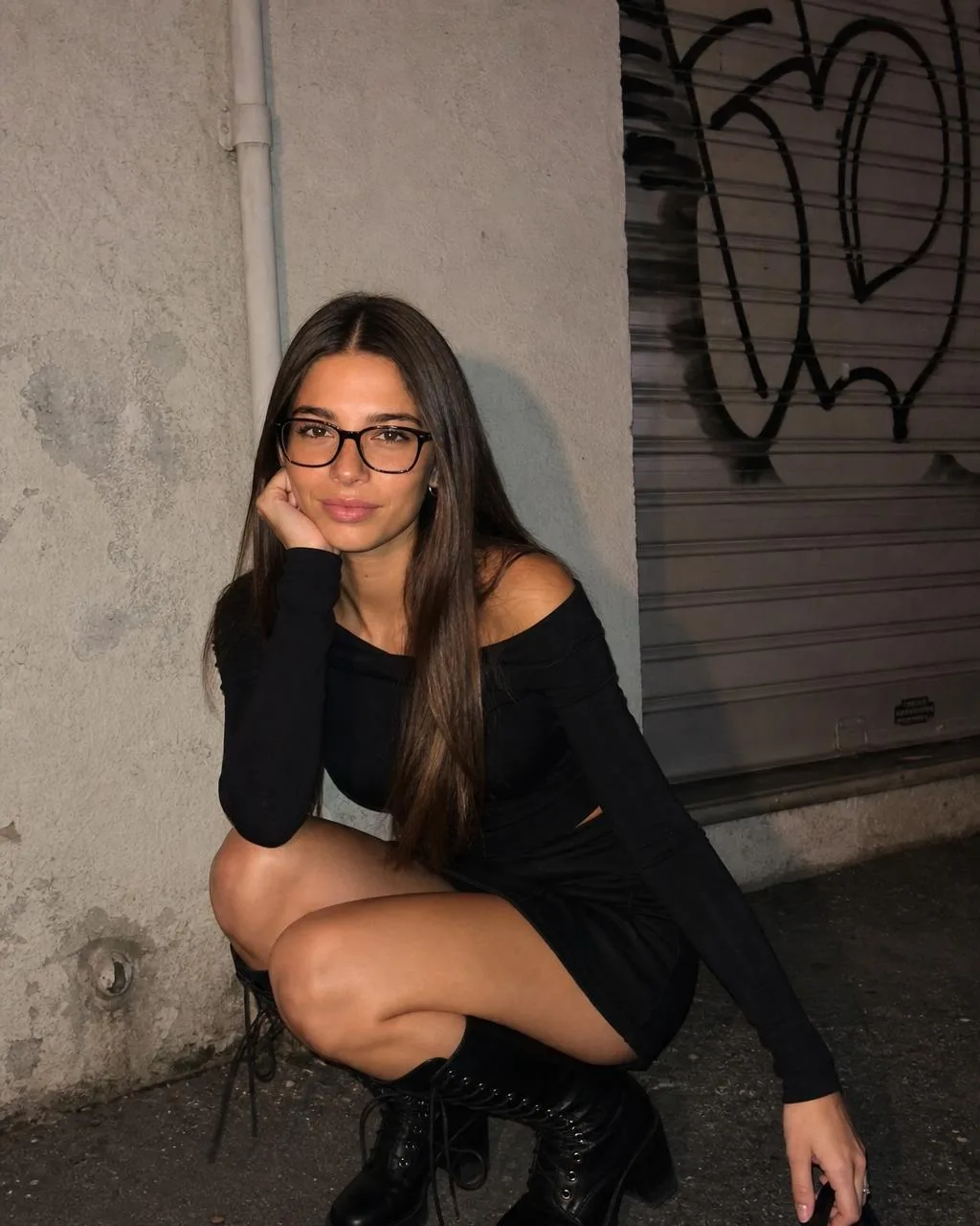 Cladegrove generated photo of the saved character crouching against a graffiti shutter on a night street, off-shoulder black top, black mini skirt, glasses and lace-up heeled boots, produced from one short prompt and one credit